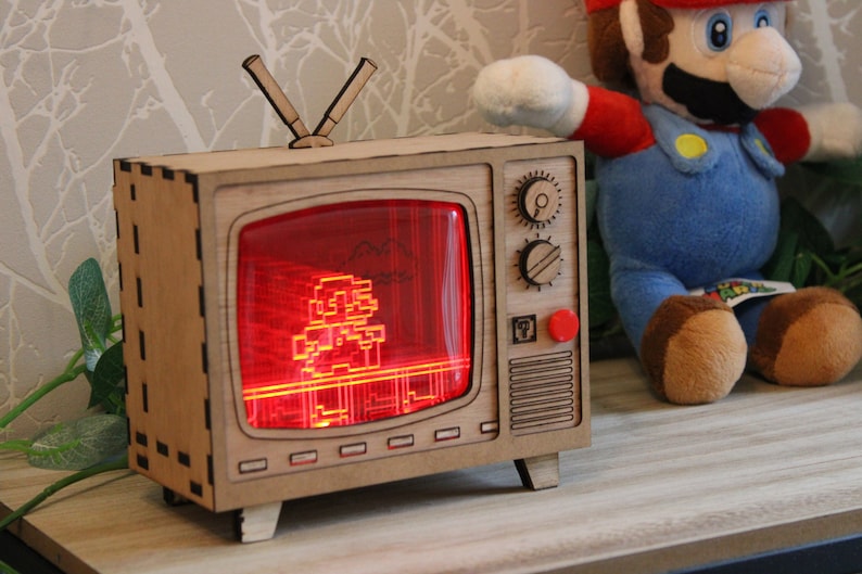 May include: A wooden retro-style television with a red screen displaying a pixelated image of a video game character. The TV has knobs, a red button, and an antenna. A plush Mario doll is in the background.