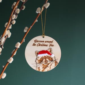 Raccoon Christmas tree ornament hanging from pussy willow branch. The ornament is engraved, laser cut wood hand finished with velvet accents. It reads “Raccoon around the Christmas Tree” and is displayed against a dark green background.