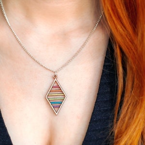 Woman wearing pendant necklace. Pendant made from wood and has sequential rainbow flag colours. It is worn on a silver plated chain by a woman with long, red hair.