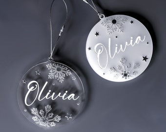 May include: Two clear acrylic Christmas baubles with the name "Olivia" engraved on them. Each bauble features a snowflake design and small stars.