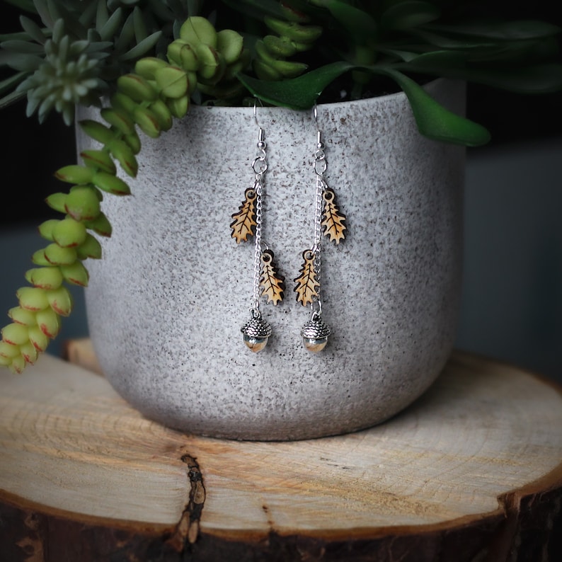 Nature inspired earrings dangling from the edge of a grey stone plant pot. The earrings feature wooden oak leaves and silver acorns.