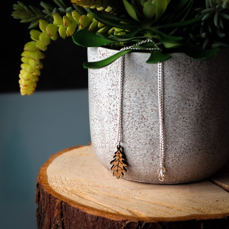 Wooden oak leaf pendant necklace dangling from plant leaves that overhang a pot. The pot is grey stone and sits on top of a log slice.