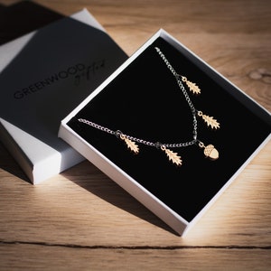 May include: A silver chain necklace with a wooden acorn charm and several wooden leaf charms. The necklace is in a white box with a black interior and the words "GREENWOOD gifted" printed on the box.