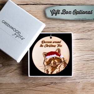 Racoon Christmas decoration in an optional gift box. The tree ornament sits on a black pillow inside a square, white gift box. The box is displayed on wood with the lid off and to the side.