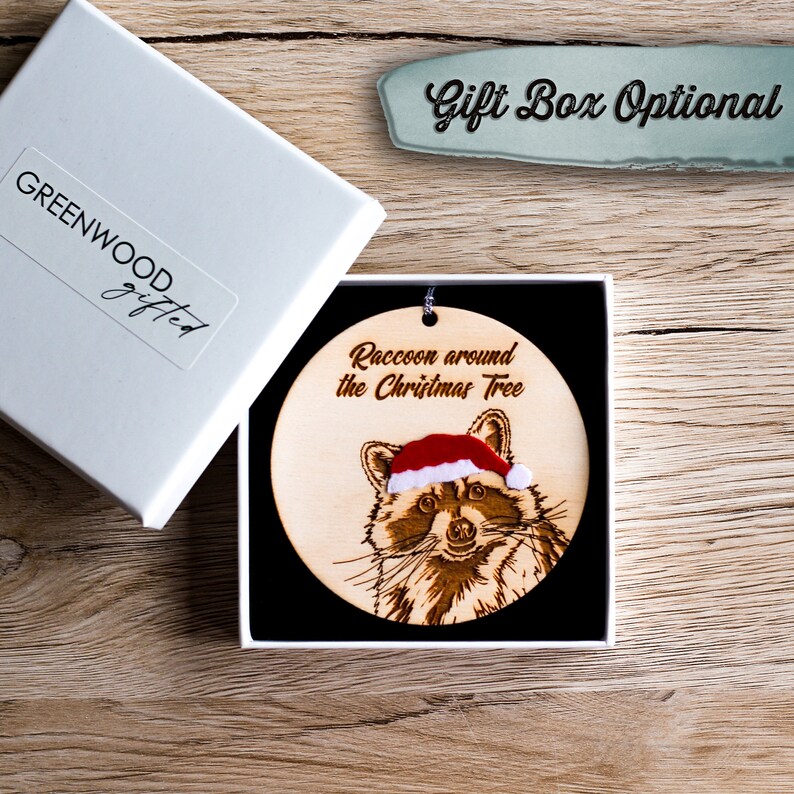 Racoon Christmas decoration in an optional gift box. The tree ornament sits on a black pillow inside a square, white gift box. The box is displayed on wood with the lid off and to the side.