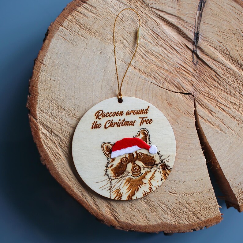 Raccoon tree decoration displayed on a log slice. The raccoon is engraved with fine detail onto wood. It wears a Santa hat with a velvet touch finish. It also has a Christmas themed pun.