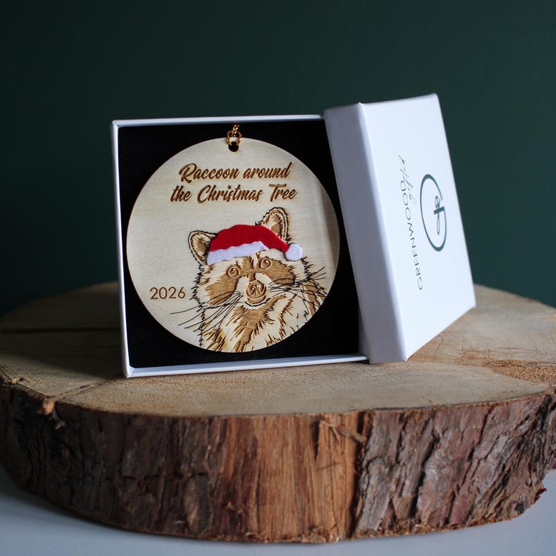 The 2026 Raccoon Christmas Ornament variant displayed upright in a gift box on a log slice against a dark green background. The gift box is square and white with greenwood gifted branding, the ornament is inside on a black pillow. It has been personalised with the year as a keepsake gift.