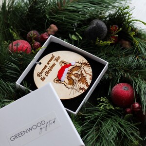 Raccoon Christmas decoction inside a gift box on a bed of Christmas foliage. The decoration features original, engraved wood with velvet accents for a mixed texture finish.