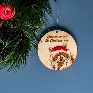 Raccoon Christmas tree ornament displayed next to a Christmas wreath on a blue background. The engraved wooden ornament reads “Raccoon around the Christmas tree” and has a velvet Santa hat.