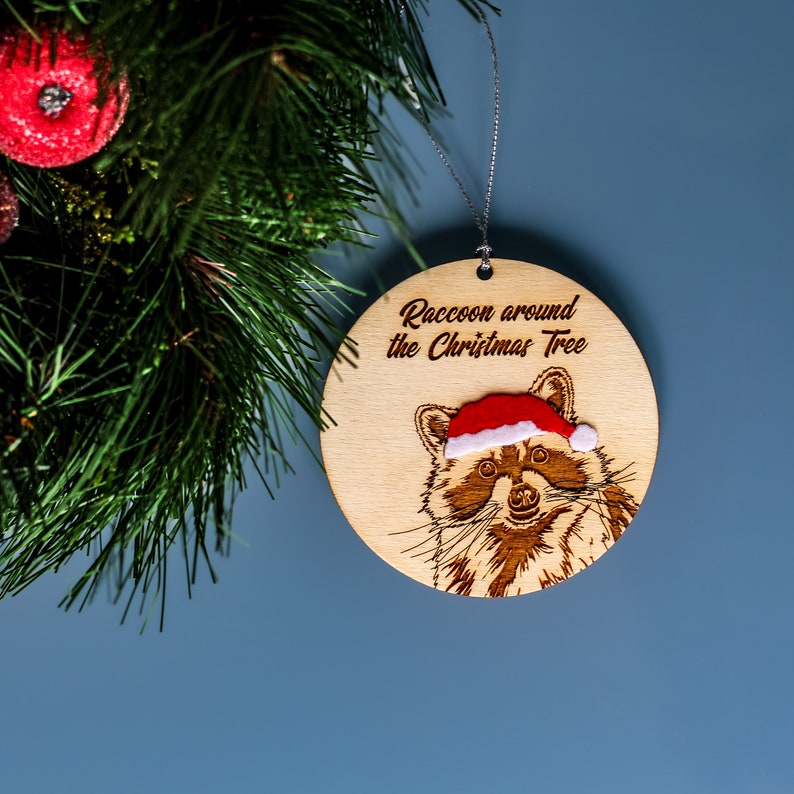 Raccoon Christmas tree ornament displayed next to a Christmas wreath on a blue background. The engraved wooden ornament reads “Raccoon around the Christmas tree” and has a velvet Santa hat.