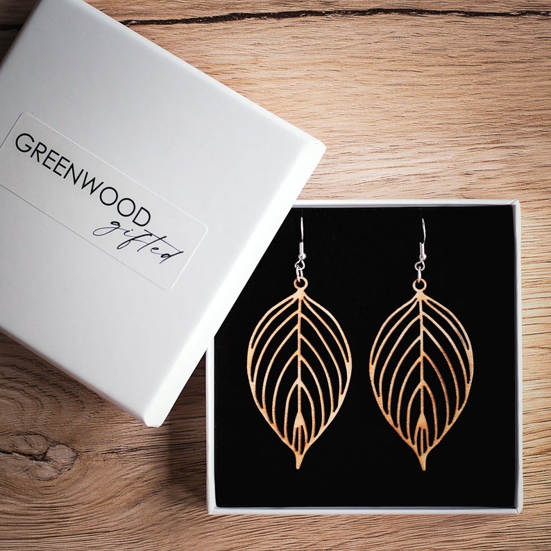 Earrings in a gift box. The gift box is white and square with a black foam pillow that the earrings are hung from. The earrings are leaf shaped, wooden and light in colour.