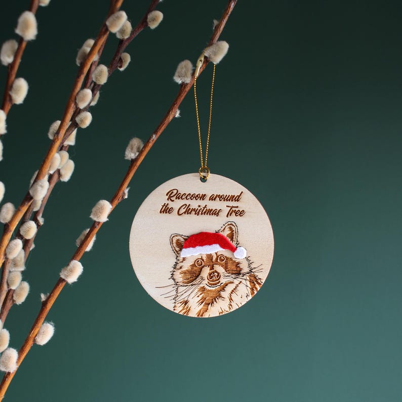 Raccoon Christmas tree ornament hanging from pussy willow branch. The ornament is engraved, laser cut wood hand finished with velvet accents. It reads “Raccoon around the Christmas Tree” and is displayed against a dark green background.