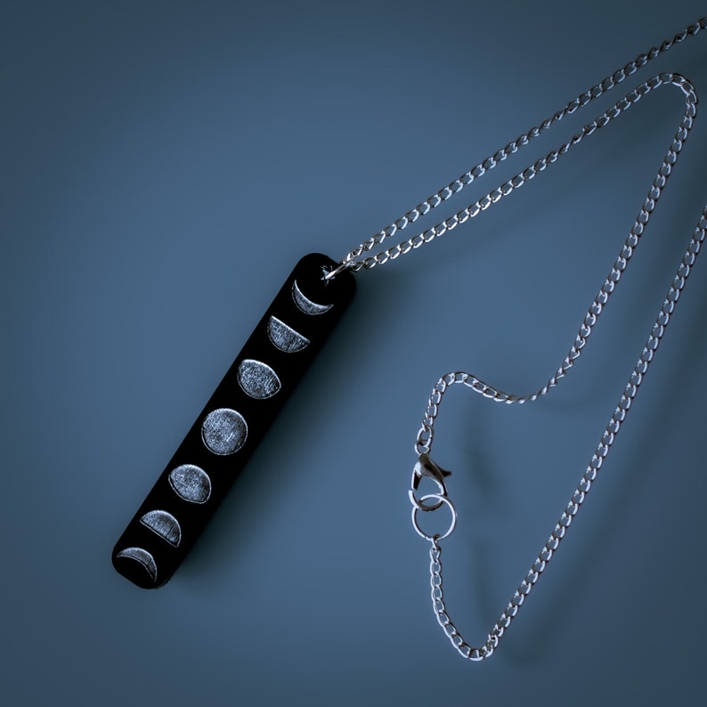 Pendant necklace on deep blue, grey background. Pendant is black with grey engravings of moon cycle stages. Chain is a thin, silver plated curb chain with a lobster clasp and large link ring for easy fastening.