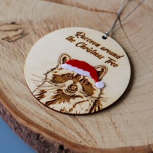 Side view of raccoon ornament. The velvet touch hat is layered on top of the wood. The wood is lower and darker where the text and drawing have been engraved.