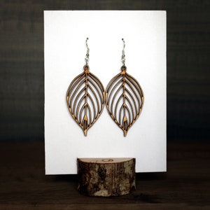 Earrings hung from a white plate that is slotted into a wooden log slice to keep upright. The earrings are made from wood attached to silver hooks. They are leaf shaped and warm and light in colour.