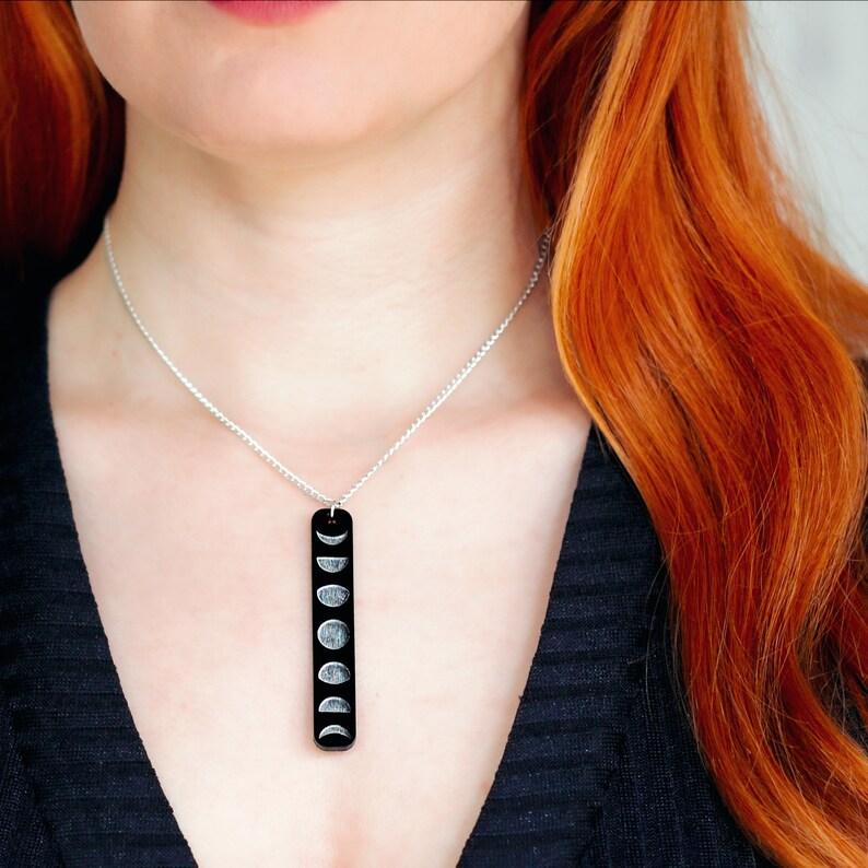Woman wearing necklace with thin, rectangular black pendant dangling from silver plated chain. Pendant is made from gloss black acrylic and engraved with moon phase design. Woman has long, red hair.