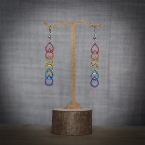Hand painted wooden earrings dangling from a gold bar that extends from a log slice. The earrings have an original, linked raindrop design with colourful metallic rainbow accents.