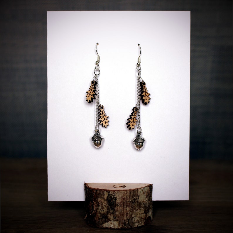Oak leaf and acorn long dangly earrings displayed on a white earring card which is inserted into a wooden log slice so that it stands upright.