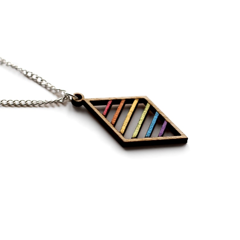 Close up on white background of wooden pendant on a silver plated chain. Pendant is a wooden diamond frame with six floating bars set within that are painted sequentially in rainbow colours.