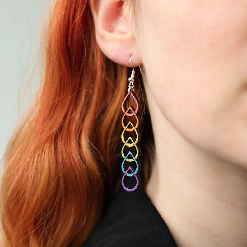 Woman wearing hand painted wooden earrings. The earrings are lightweight with a long dangle to them. They have a silver ear hook and a rainbow pride coloured, linked teardrop design.