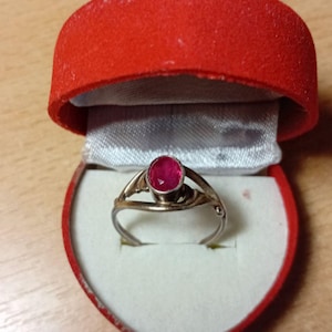 May include: A gold and silver ring featuring a large, round, red gemstone, displayed in a red heart-shaped box with a white satin lining. The ring's band has an ornate design.