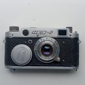 Puede incluir: Una cámara FED-2 vintage con un cuerpo negro texturizado y detalles plateados. La cámara presenta una lente con ajustes de apertura, una tapa de lente plateada y el texto "FED-2" en escritura cirílica. Un ejemplo clásico de equipo fotográfico de la era soviética.