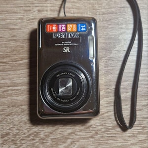 Puede incluir: Cámara digital Pentax plateada con correa de muñeca negra. La cámara tiene una lente con el texto "Pentax Lens" y "Optical 5x Zoom". La cámara también tiene el texto "Pentax" y "SR" en la parte delantera.