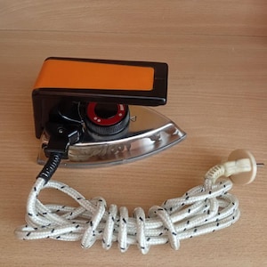 Puede incluir: Una plancha eléctrica vintage con suela plateada, mango negro y acento naranja. La plancha tiene un dial rojo y un cable blanco con detalles negros. El cable está enrollado.