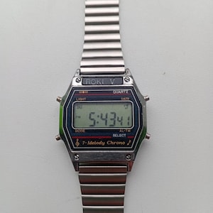 May include: A silver-toned digital watch with a rectangular face and a matching metal band. The watch face displays the time as 5:43:41, with the text "ROKI V" at the top. Additional text includes "QUARTZ", "LIGHT", "DATE", "MODE", "AL-TM", "SELECT", and "7-Melody Chrono".