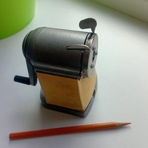 Puede incluir: Un sacapuntas vintage de color plateado con una base de madera y una manivela negra. Un lápiz naranja se encuentra horizontalmente frente al sacapuntas. El sacapuntas tiene una palanca y una pequeña abertura para el lápiz.