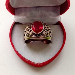 Könnte beinhalten: Ein goldfarbener Ring mit einem großen, ovalen, roten Edelstein wird in einer roten herzförmigen Schatulle präsentiert. Der Ring weist aufwendige Filigranarbeiten um den Edelstein auf. Die Schatulle ist mit hellgrauem Stoff ausgekleidet.