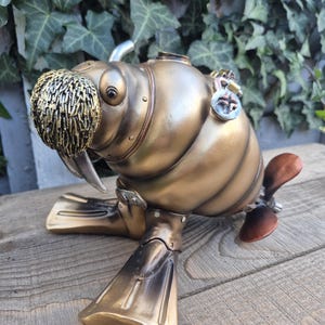 May include: A steampunk walrus sculpture in metallic gold. The walrus has a textured, gold-coloured moustache, large tusks, and flipper-like feet. Details include rivets, a propeller, and a circular emblem. The sculpture is on a wooden surface.