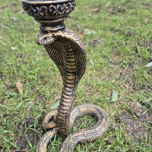 May include: A cobra-shaped candle holder featuring a coiled snake base with textured scales in a bronze and black finish. The upright cobra supports a circular, ornate candle cup at the top, designed with intricate scrollwork along the outer rim.