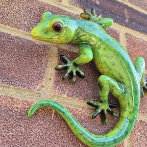 May include: A glossy green lizard figurine with a marbled texture and dark brown eyes. The lizard has a long, curved tail and four feet with distinct toes, positioned as if climbing. The surface of the lizard is smooth and reflective.