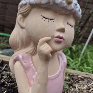 May include: A ceramic planter shaped like a girl's bust with closed eyes and a flower crown. The planter is wearing a pink dress and has a light brown skin tone.