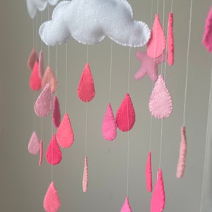 Puede incluir: Una nube de fieltro blanco con gotas de lluvia de fieltro rosa colgando de ella. La nube es la pieza central de un móvil con una estrella de fieltro rosa.