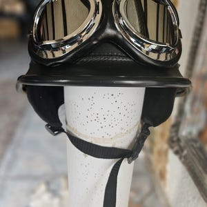 Può includere: Un casco da moto in pelle nera dotato di una visiera anteriore prominente e di un paio di occhiali protettivi con montatura cromata e lenti scure. Il casco include un sottogola nero con chiusura a fibbia in plastica per una tenuta sicura.