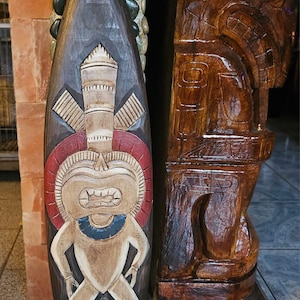 May include: A carved wooden tiki mask with a dark brown finish. The mask features a central figure with a red and white circular design, a blue collar, and a detailed face. A second, larger wooden tiki statue is visible to the right.