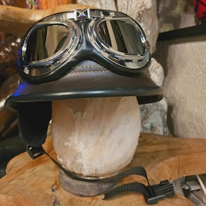 May include: A brown leather motorcycle helmet featuring a black brim and integrated chrome-rimmed goggles with dark reflective lenses. The helmet includes a black chin strap with a plastic buckle and side ear flaps, providing a classic vintage aesthetic.