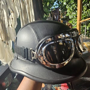 Può includere: Un casco da moto nero opaco dal design arrotondato, dotato di un paio di occhiali con montatura cromata e lenti scure, fissati da un cinturino elastico nero che avvolge l'esterno del casco.