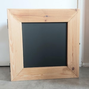 May include: A square chalkboard with a natural wood frame. The frame is made of light-colored wood with visible grain and knots. The chalkboard surface is black and fills the center of the frame.