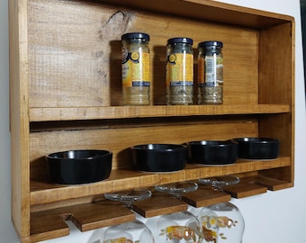 May include: A wall-mounted wooden shelf with a natural brown finish featuring two tiers. The top tier holds three glass spice jars with yellow labels. The bottom tier holds four small black bowls and three hanging wine glasses with graphic prints.