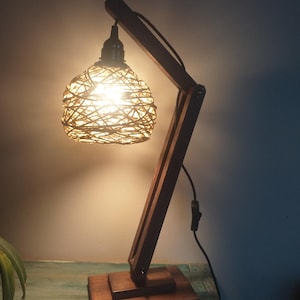 Op de afbeelding: Een houten tafellamp met een bruine houten voet en een geweven lampenkap. De lamp is aan en het licht schijnt door de kap.