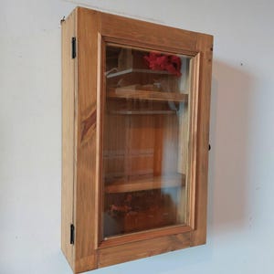 May include: A wooden wall-mounted cabinet with a glass door. The cabinet is made of light brown wood and has a rectangular shape. Inside, there are shelves and a red cloth. The cabinet is mounted on a white wall.