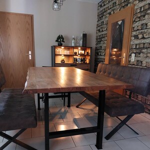 May include: A wooden dining table with a live edge top and black metal legs. The table is surrounded by two brown leather benches with black metal legs. The table is set in a dining room with a brick wall and a wooden cabinet with the word "HOME" on it.