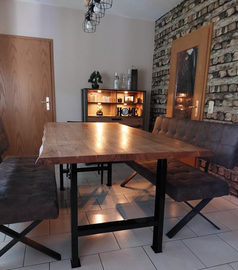 May include: A wooden dining table with a live edge top and black metal legs. The table is surrounded by two brown leather benches with black metal legs. The table is set in a dining room with a brick wall and a wooden cabinet with the word "HOME" on it.