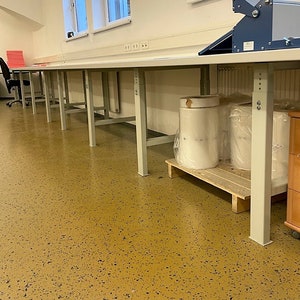 May include: A long, white work table with metal legs stands in a room with a speckled floor. The table is empty except for a few rolls of clear plastic wrap on a wooden pallet.