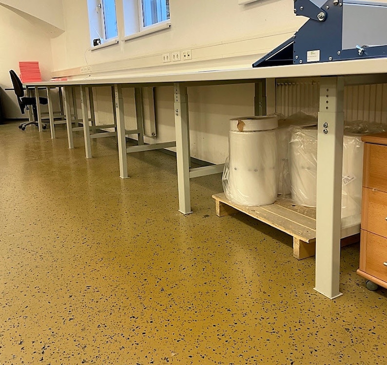 May include: A long, white work table with metal legs stands in a room with a speckled floor. The table is empty except for a few rolls of clear plastic wrap on a wooden pallet.
