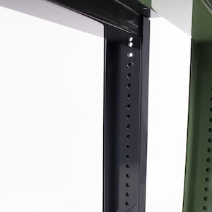 May include: Close-up of a black metal rack with a row of holes for mounting equipment. The rack is part of a larger metal shelving unit with a green frame.