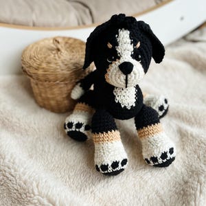 May include: A crocheted stuffed dog toy with black, white, and tan fur. The dog has black eyes and a black nose. The dog is sitting on a white fluffy blanket.